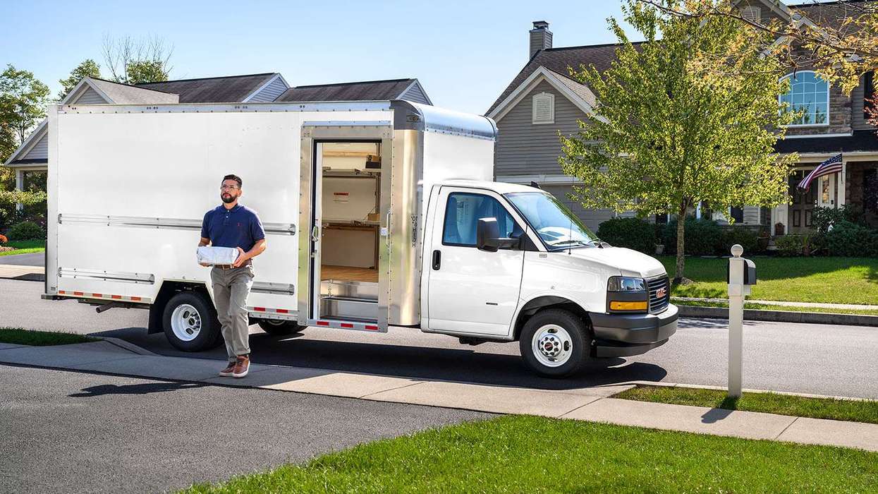 A delivery driver carries a package walking away from a white box truck up a driveway.