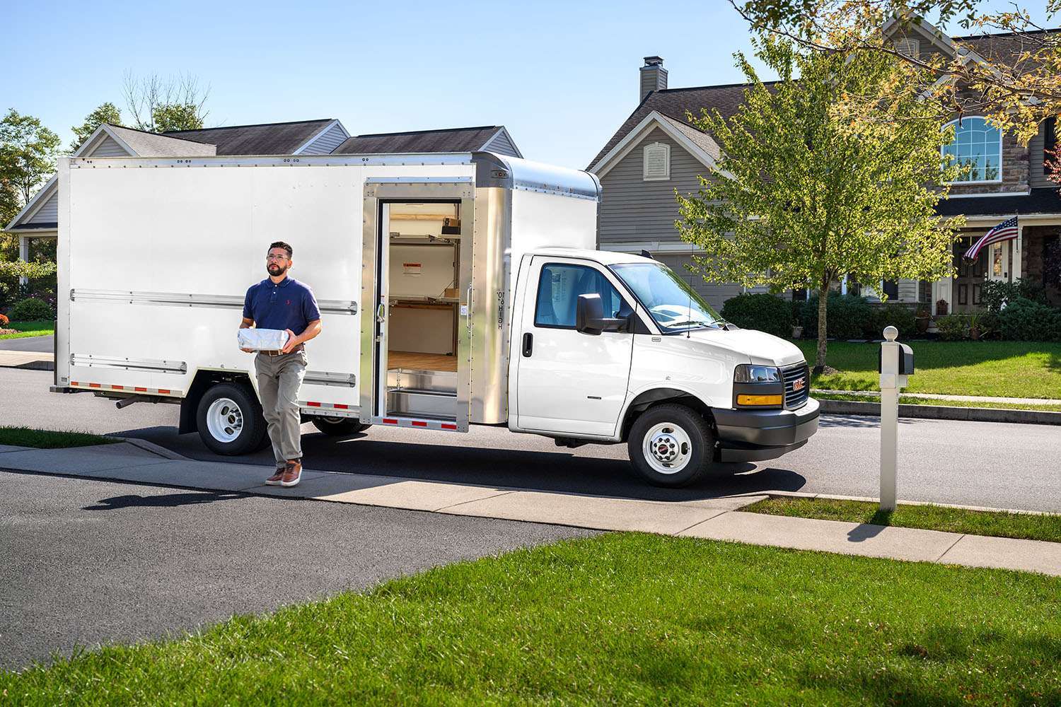 A delivery driver carries a package walking away from a white box truck up a driveway.
