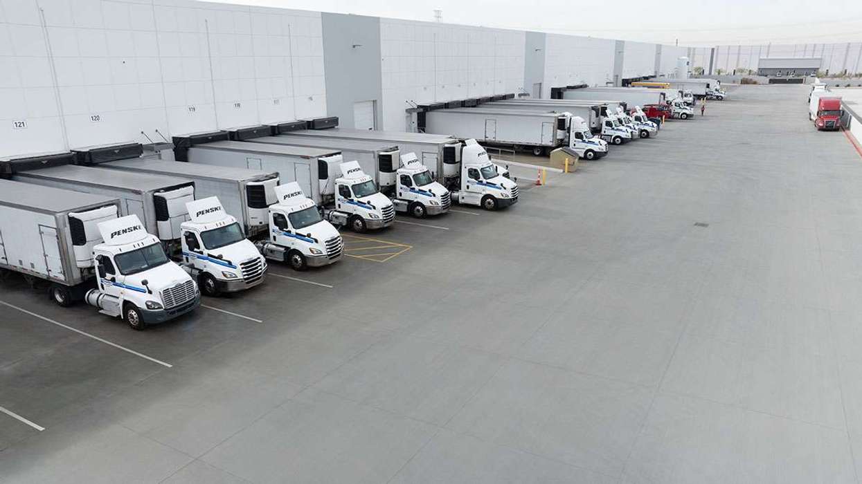 A fleet of white Penske semi-trucks are parked in a line backed up to a big warehouse loading dock.