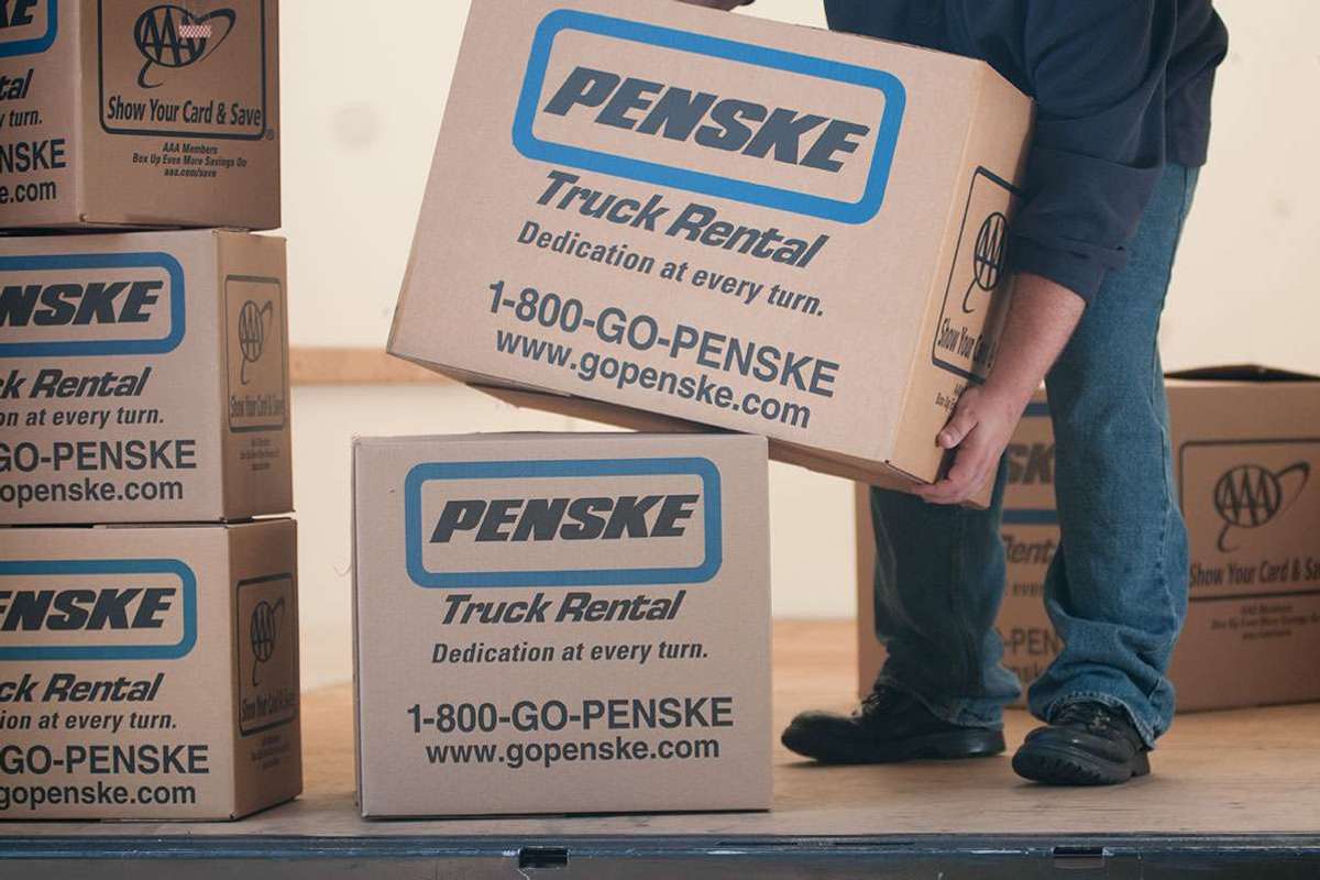 A person in blue jeans and a blue shirt bends over to lift a cardboard Penske box.