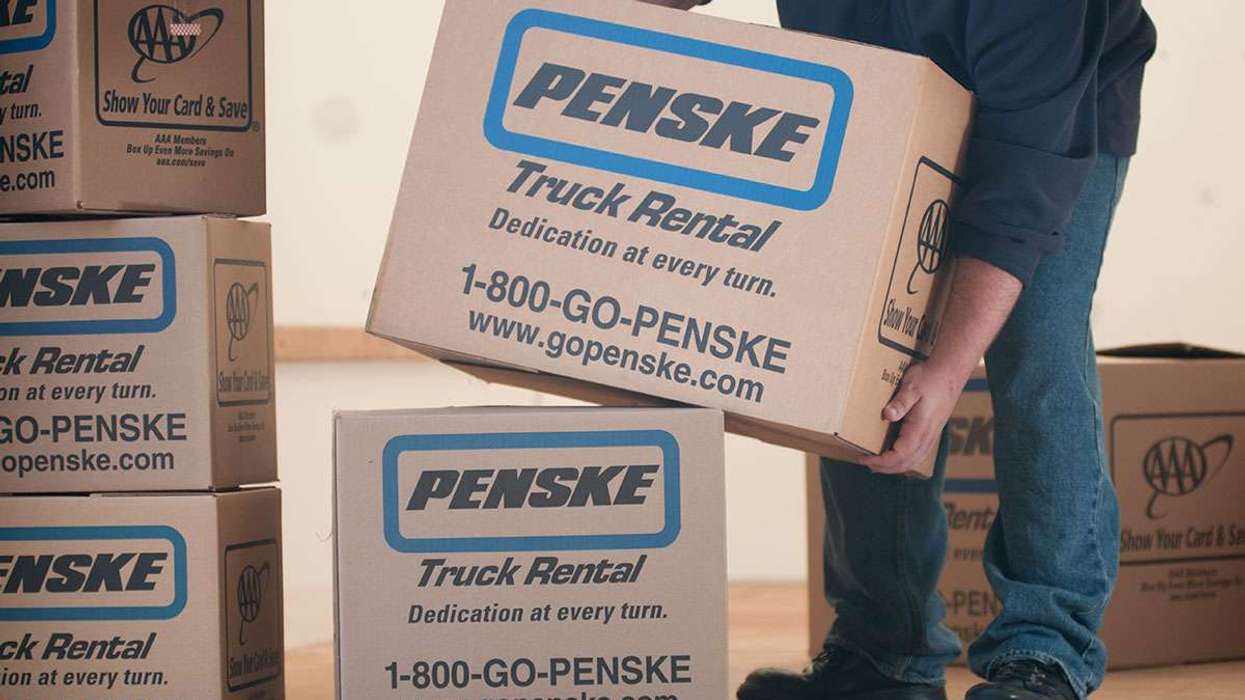 A person in blue jeans and a blue shirt bends over to lift a cardboard Penske box.