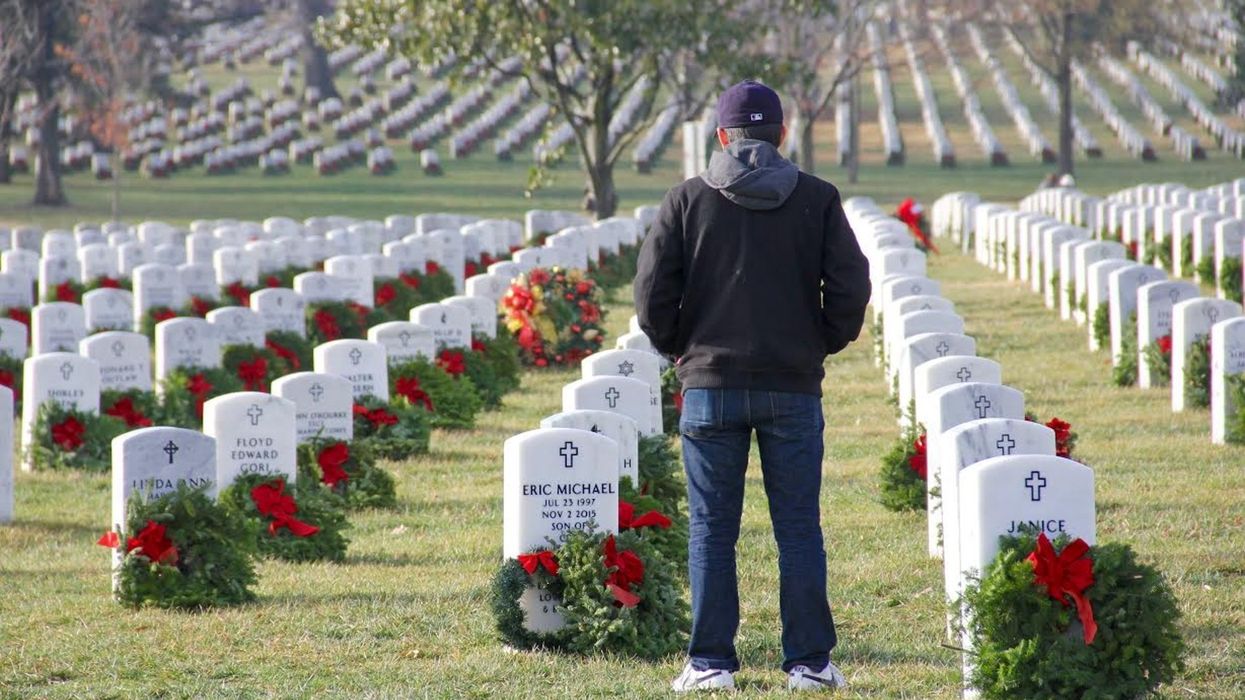 A person stands solemnly at a military cemetery surrounded by rows of wreath-adorned headstones, honoring veterans as part of Wreaths Across America.