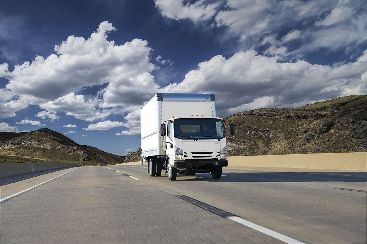 A white cabover truck drives up a road under a blue cloudy sky.