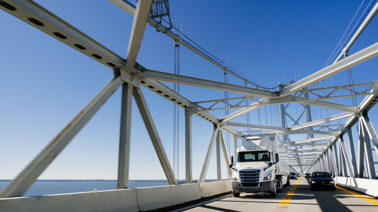 A white Penske semi-truck drives over a bridge on a sunny day.