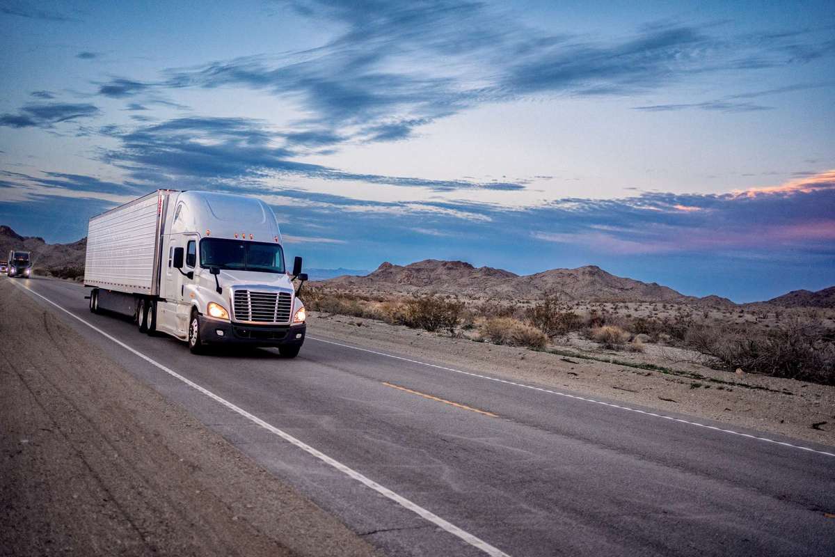 A white semi-truck drives up a desert road at sunset.