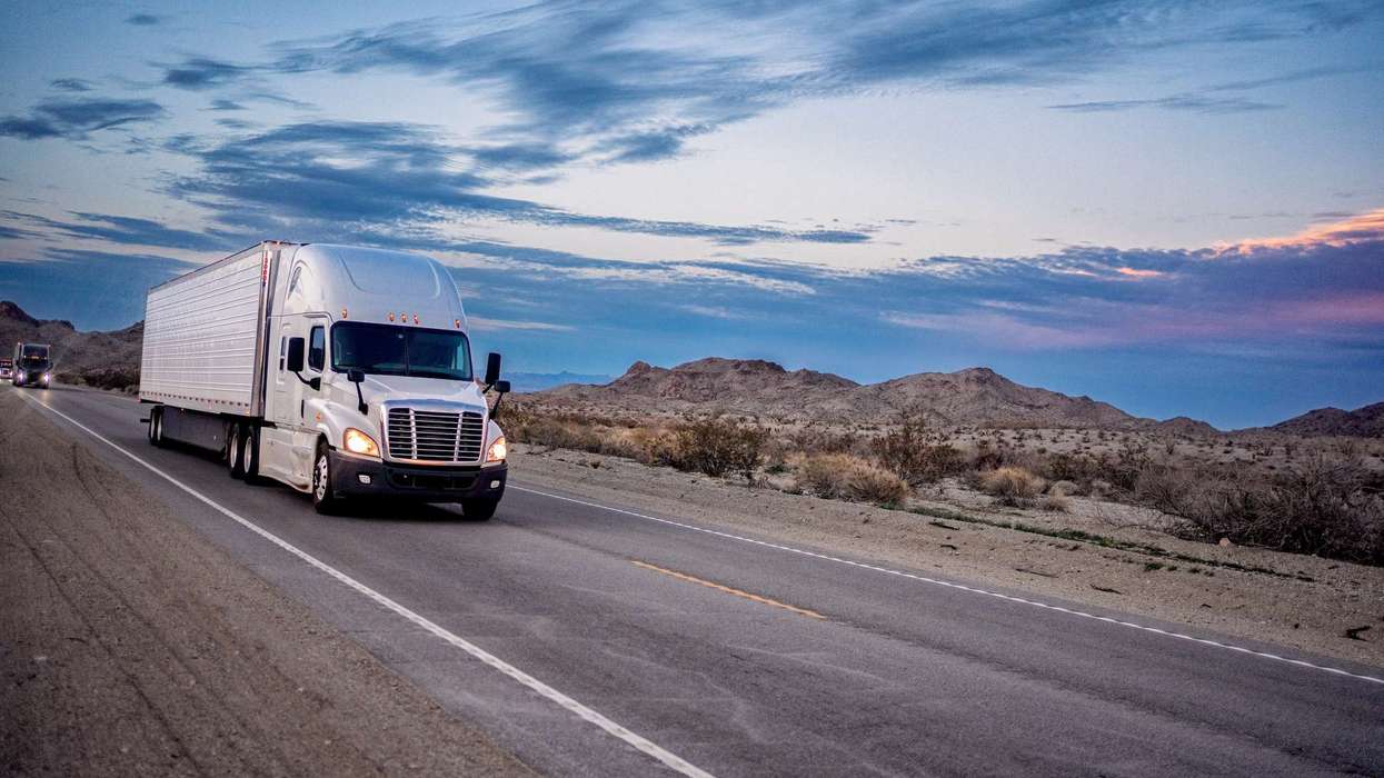 A white semi-truck drives up a desert road at sunset.