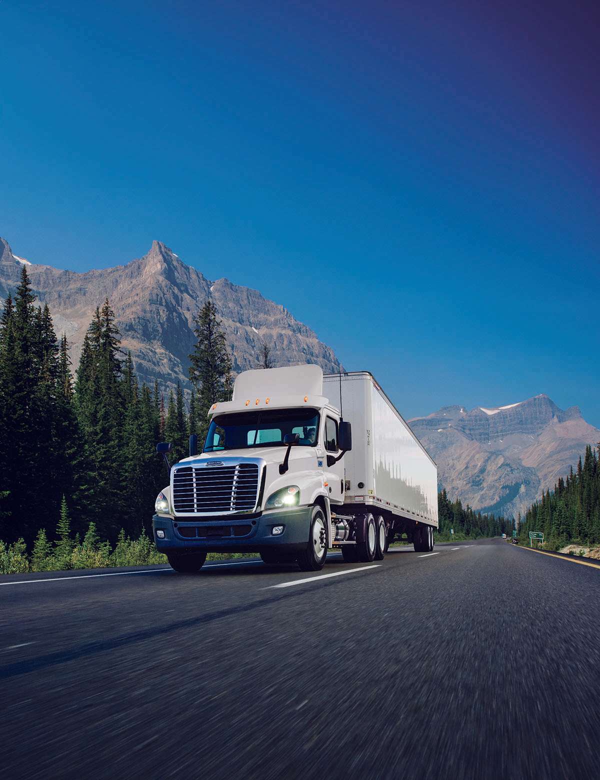 A white used Penske semi-truck drives up a road with mountains in the background.