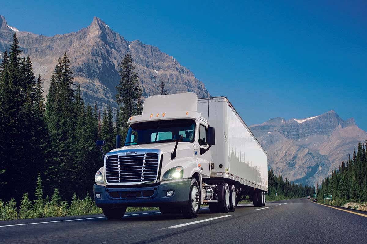 A white used Penske semi-truck drives up a road with mountains in the background.
