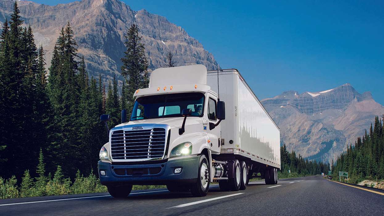 A white used Penske semi-truck drives up a road with mountains in the background.