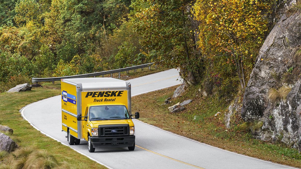 A yellow 16 foot Penske box truck drives along a curvy road surrounded by green trees.