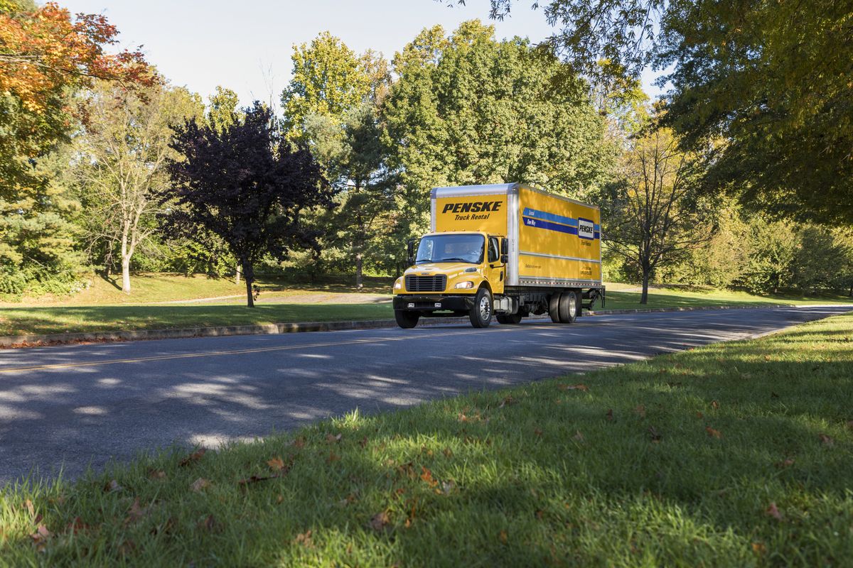 A yellow 26 foot Penske truck drives up a road.