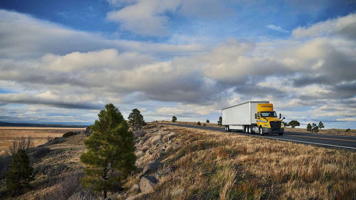 A yellow and white Penske semi-truck drives along a country road under a blue cloudy sky.