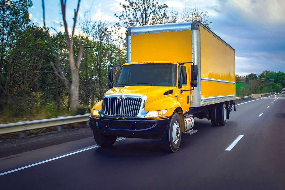 A yellow box truck drives on a freeway next to trees.