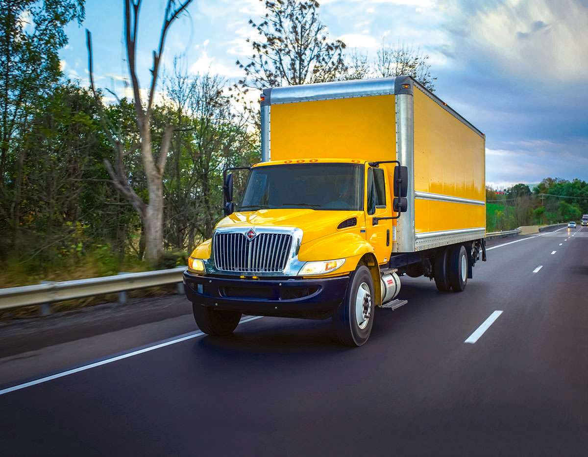 A yellow box truck drives on a freeway next to trees.