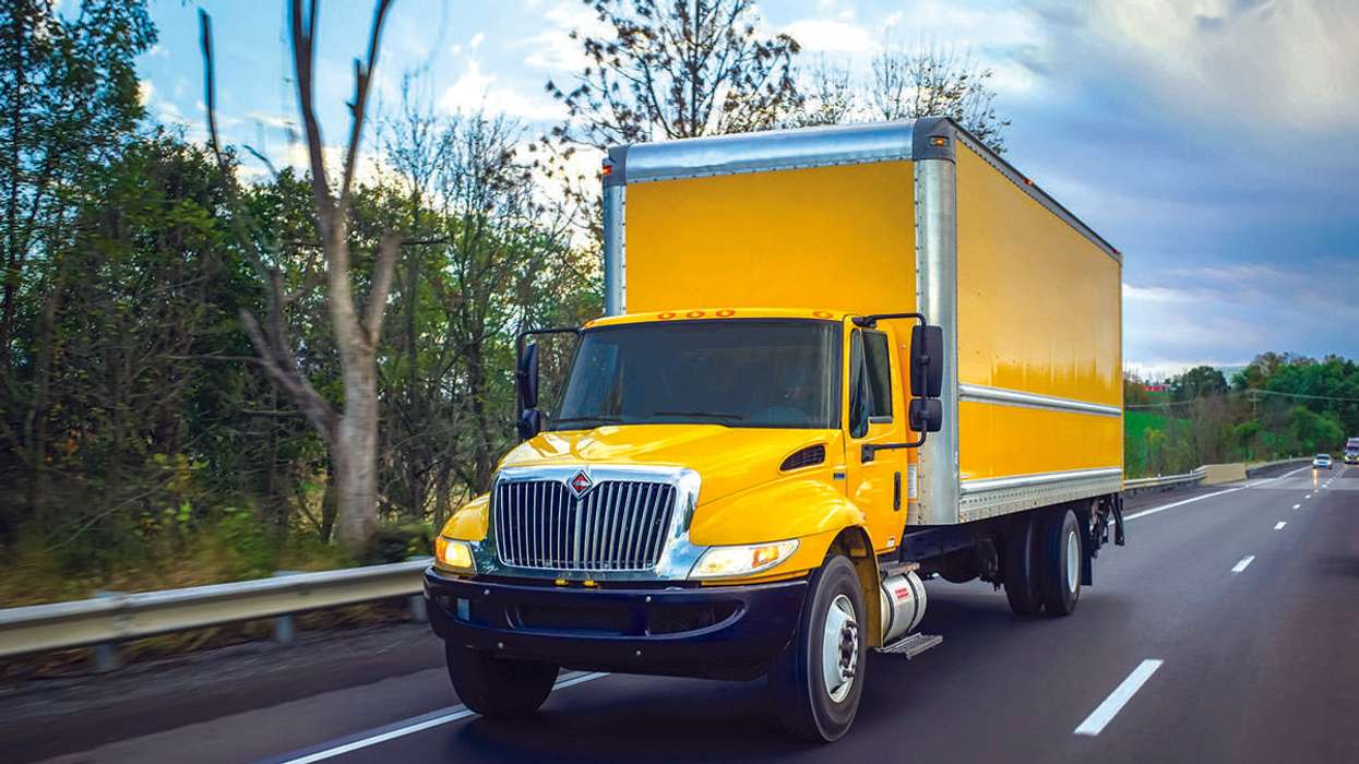 A yellow box truck drives on a freeway next to trees.