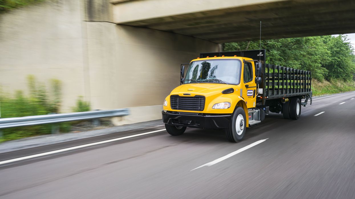 A yellow Penske flatbed truck drives on a freeway under a bridge.
