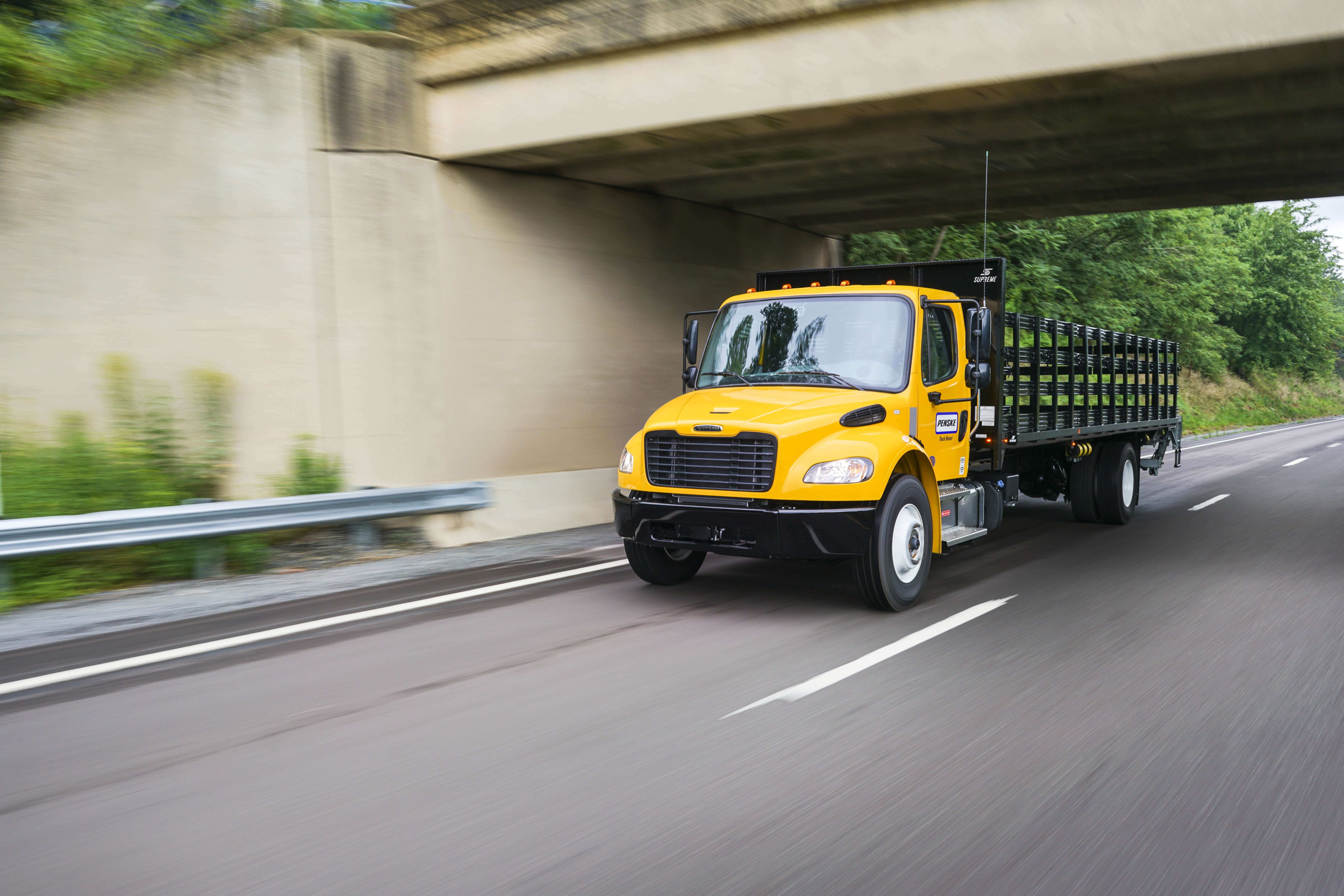 A yellow Penske flatbed truck drives on a freeway under a bridge.