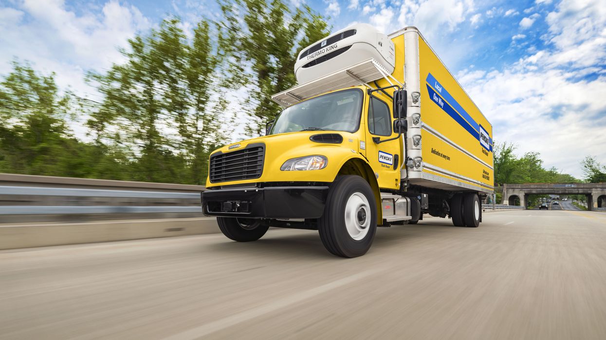 A yellow Penske refrigerated box truck drives up a freeway on a blue sky day.