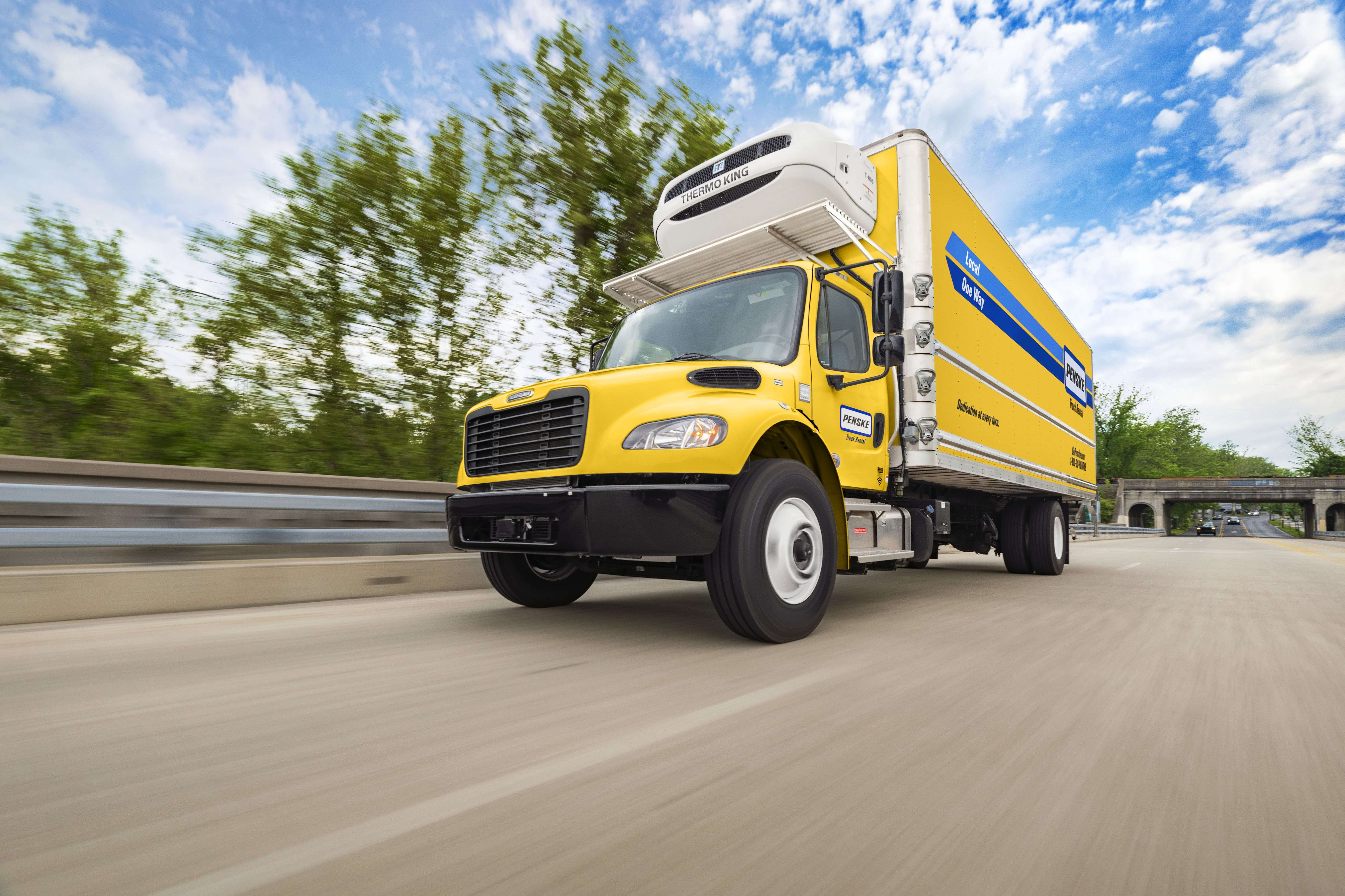 A yellow Penske refrigerated box truck drives up a freeway on a blue sky day.