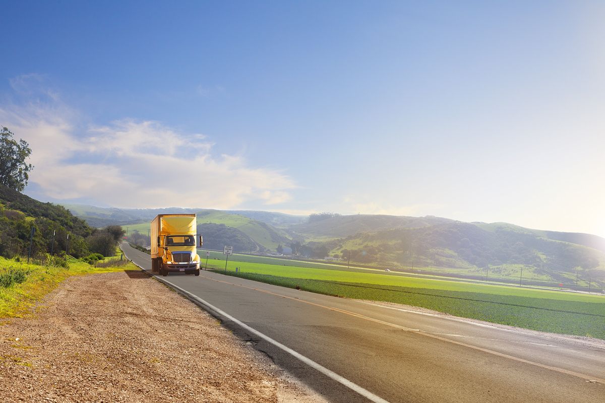 A yellow truck drives through the country on a sunny day.