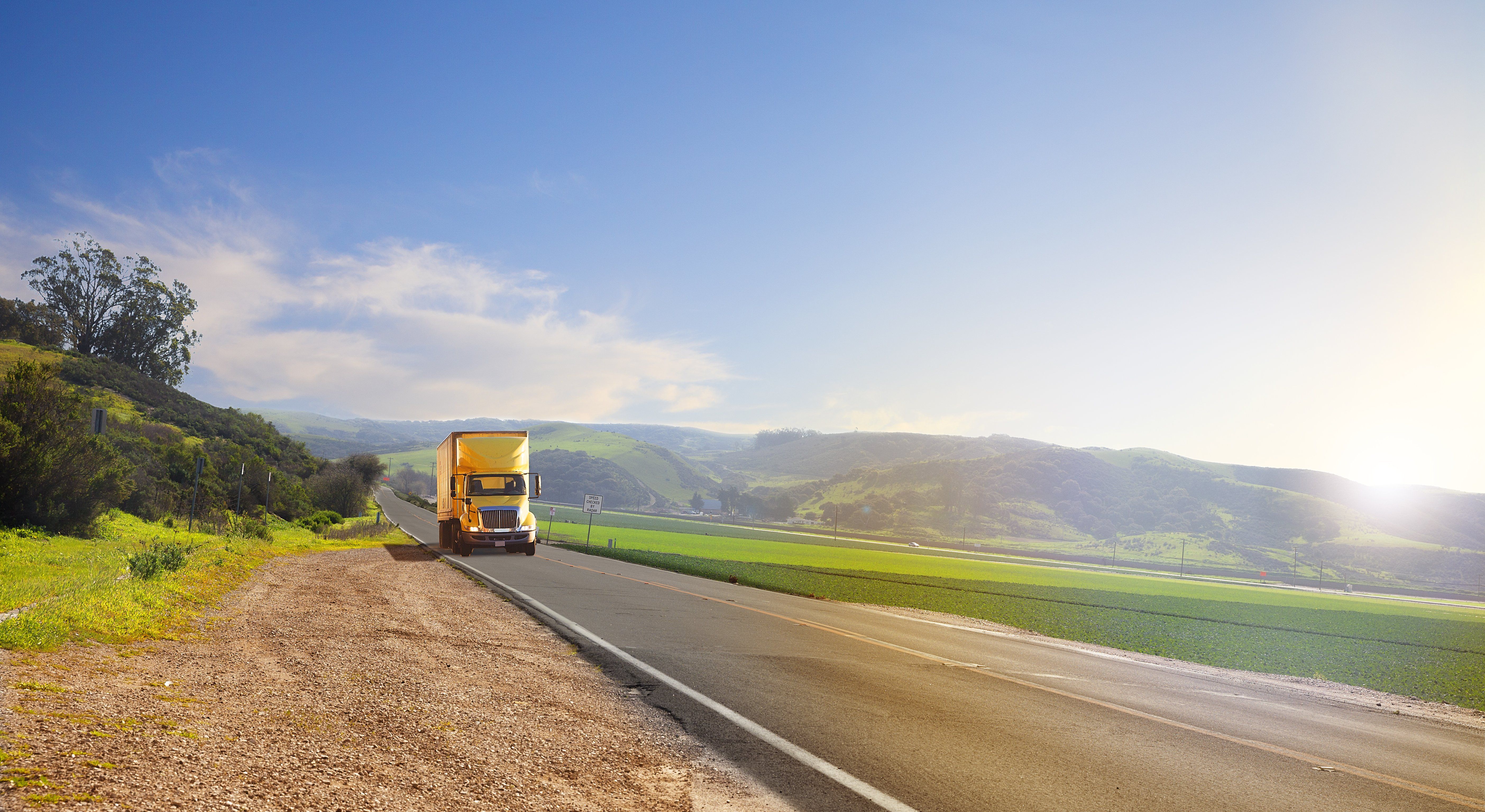 A yellow truck drives through the country on a sunny day.