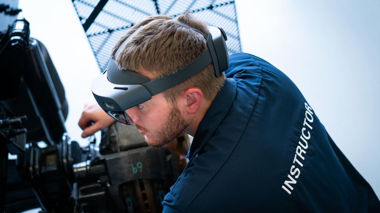 Penske instructor, wearing a blue uniform with the word instructor across the back in all caps, crouched down looking at a diesel truck engine. He is wearing mixed reality HoloLens Goggles for training purposes.
