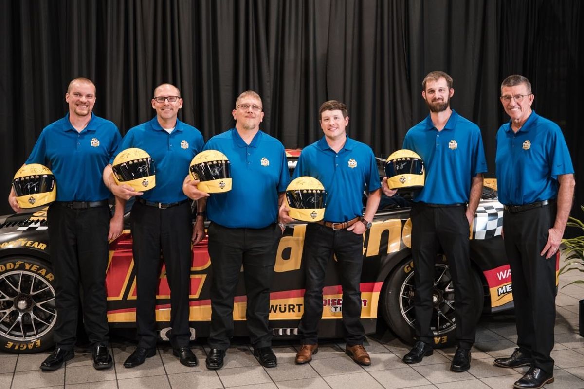 Six men posing in front of a race car holding helmets