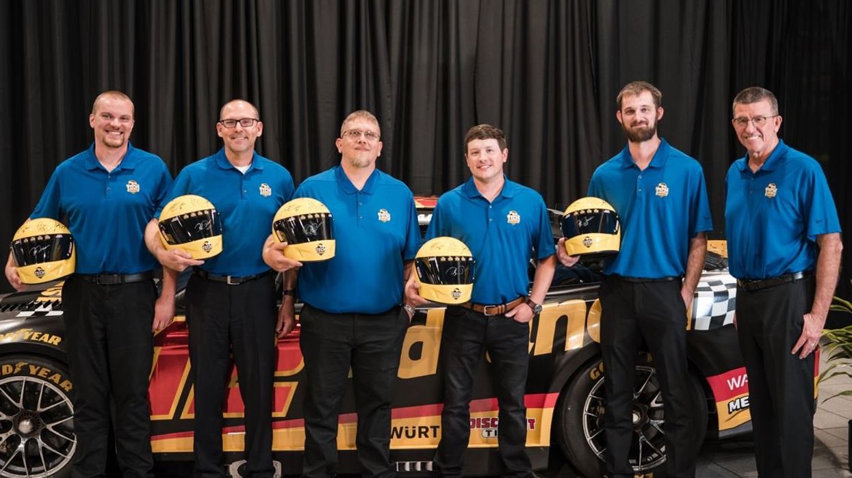 Six men posing in front of a race car holding helmets