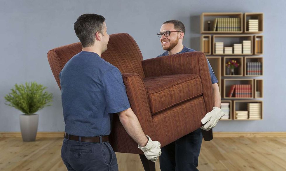 Two men wearing gloves carry a red striped chair through a living room.
