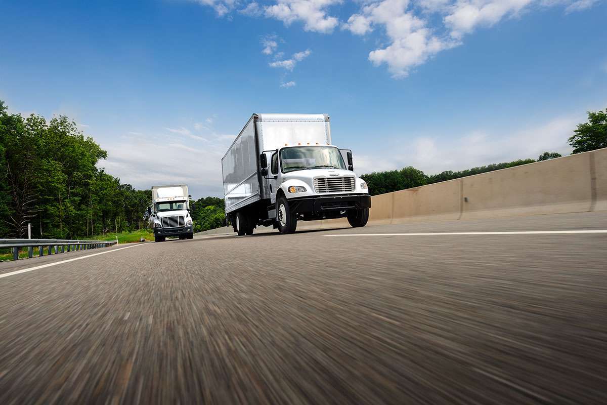 Two white trucks travel on a highway under a blue, cloudy sky.