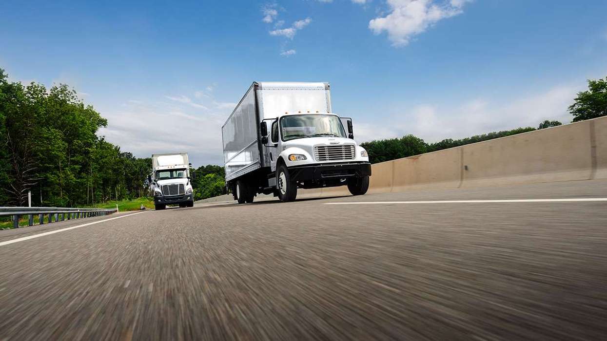 Two white trucks travel on a highway under a blue, cloudy sky.