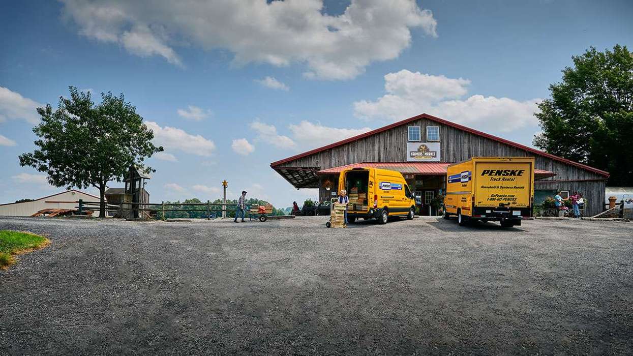 Two yellow Penske trucks unload boxes outside a brown barn farmer's market