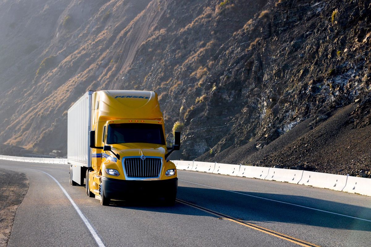 Yellow Penske tractor trailer driving down a highway past mountains